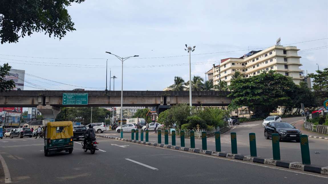 Arayidathupalam junction, Kozhikode: the concrete flyover crossing the frame with Kerala PWD direction boards, an autorickshaw and motorcycle in the foreground, bollarded median down the carriageway and the hospital block beyond
