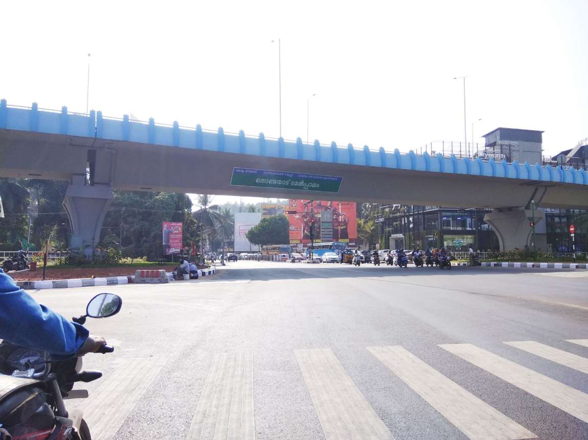 Thondayad flyover junction, Kozhikode: the blue-railed span crossing above the bypass with its Malayalam PWD name board, showrooms and a mall frontage below, and two-wheelers and cars queued along the service road