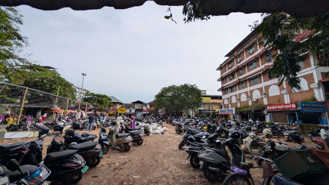 The unpaved yard behind SM Street, Kozhikode, packed edge to edge with parked scooters and motorcycles on bare earth, waste sacks heaped in the middle and shopfronts along the far side