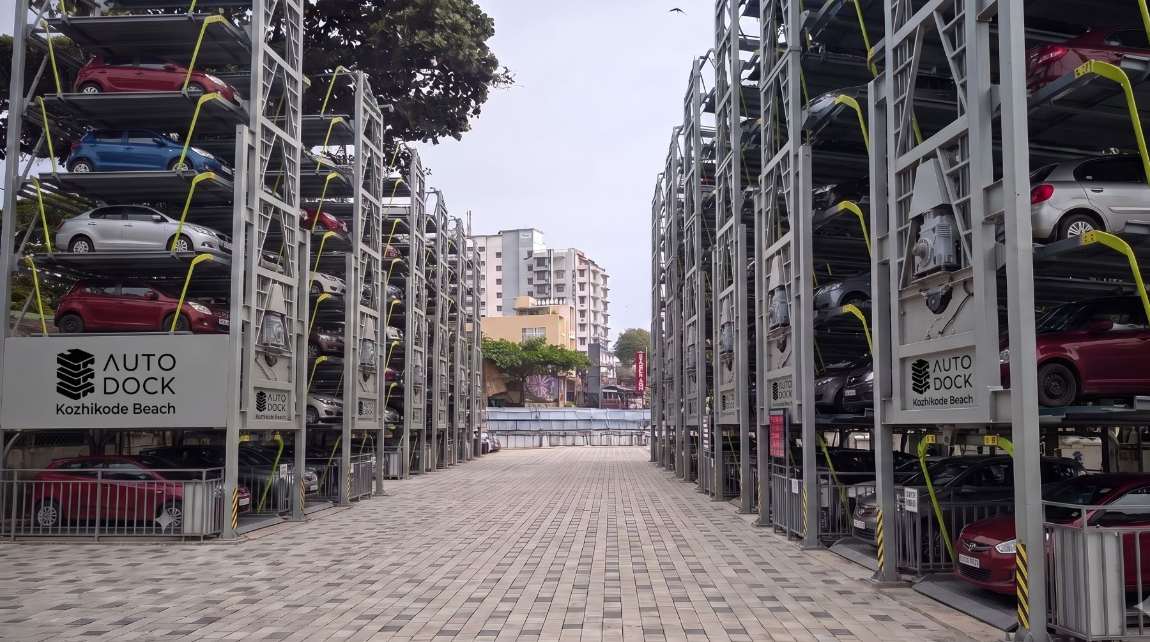 The same Kozhikode Beach plot rebuilt with rows of multi-level rotary parking towers on both sides, AutoDock Kozhikode Beach signage, and a paved block aisle down the centre