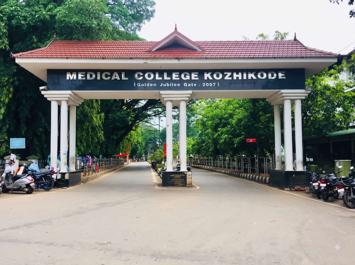 The Golden Jubilee Gate at Medical College Kozhikode: a tiled-roof gateway over the approach road with two-wheelers parked along the railings on both verges and a tree-lined avenue beyond