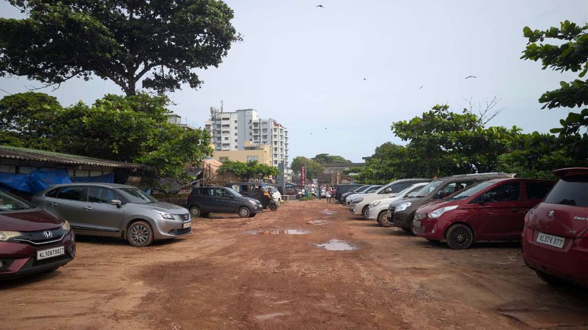 The unpaved laterite lot serving Kozhikode Beach: cars parked in two rough rows on bare earth with rain puddles down the centre and a hand-painted CAR PARKING sign at the far end